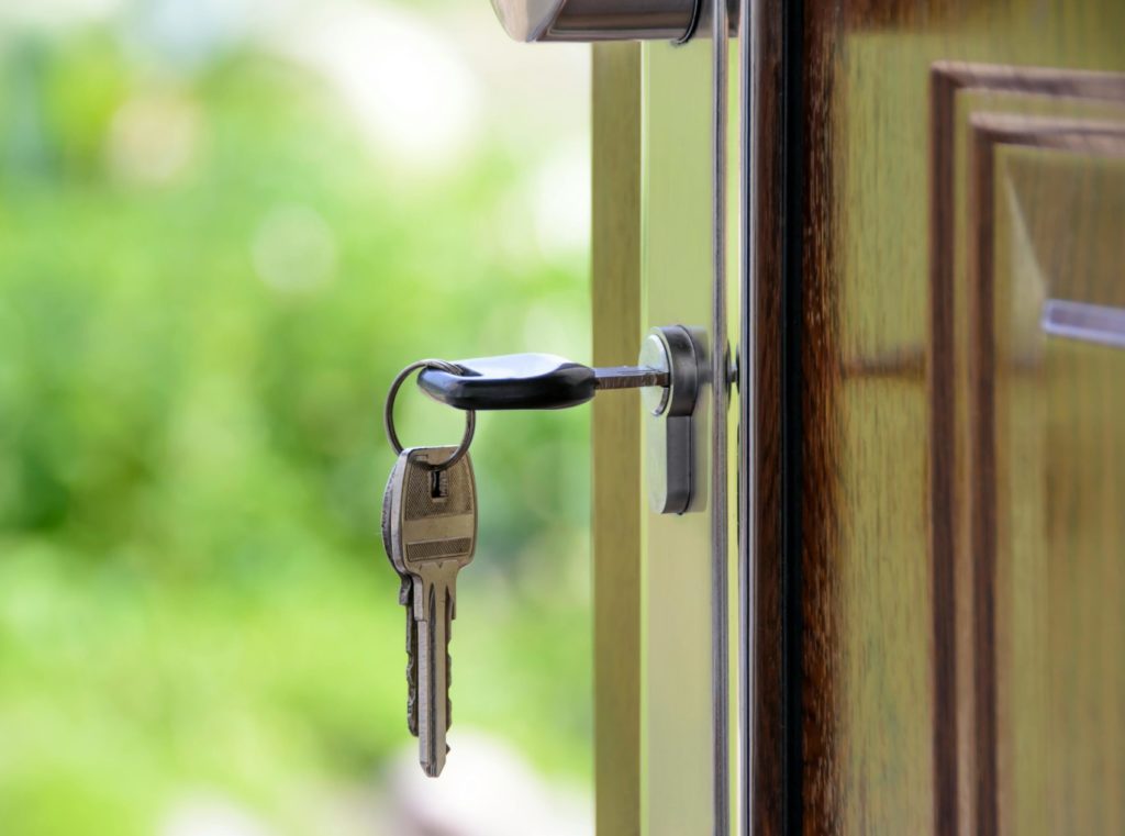 A set of keys is inserted into a door lock with a blurred natural background visible through the open doorway.