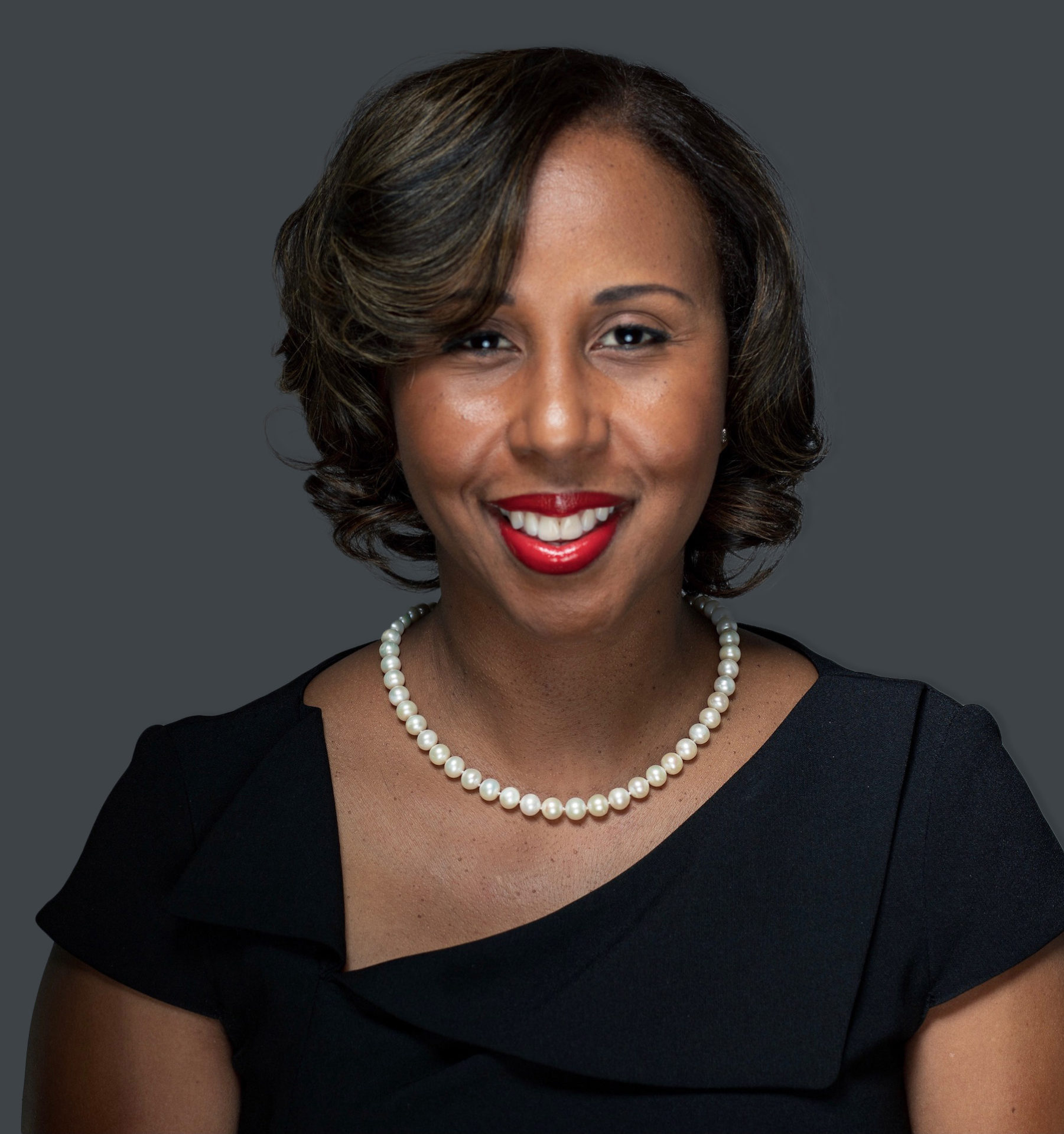 A smiling individual with pearl necklace is wearing a black outfit against a grey background.