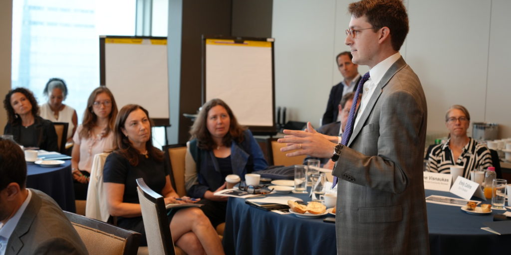 A professional meeting is taking place with a person standing and speaking while others listen attentively around a table with beverages and notepads.