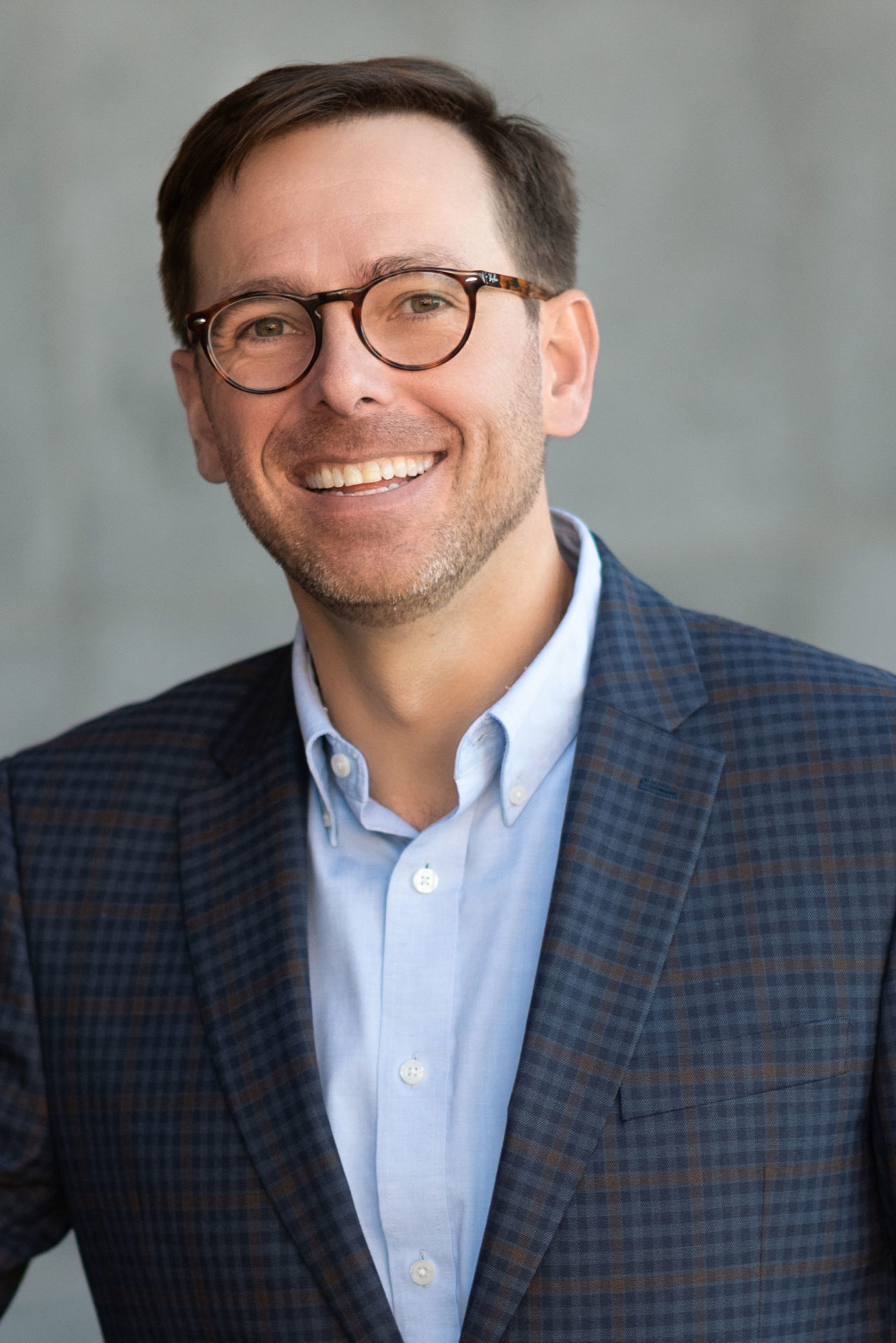 A smiling individual wearing glasses, a checkered blazer, and a blue shirt poses for a portrait.