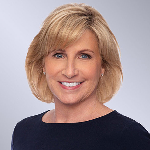 A smiling woman with short blond hair wearing a dark top poses for a professional portrait against a grey background.