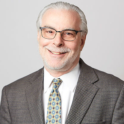 A smiling individual wearing glasses, a checked blazer, and a patterned tie poses against a plain background.