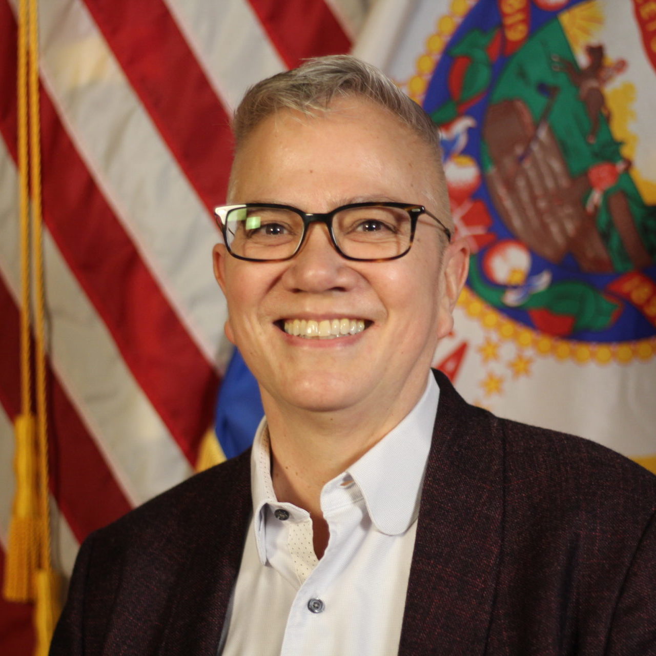 A smiling person with short hair wearing glasses, a blazer over a button-up shirt, stands in front of a ceremonial flag.