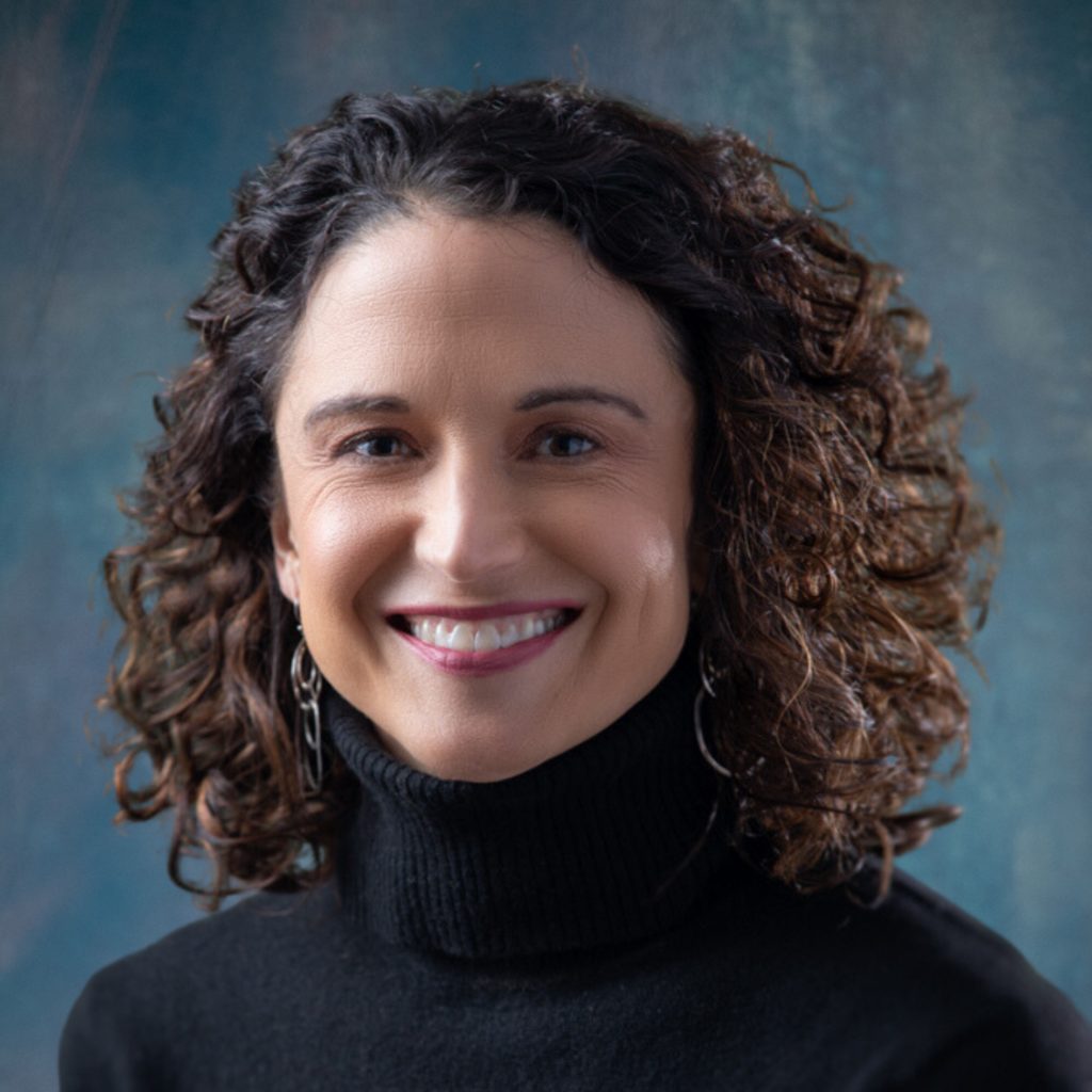 A smiling individual with curly hair is wearing a black turtleneck against a mottled blue background.