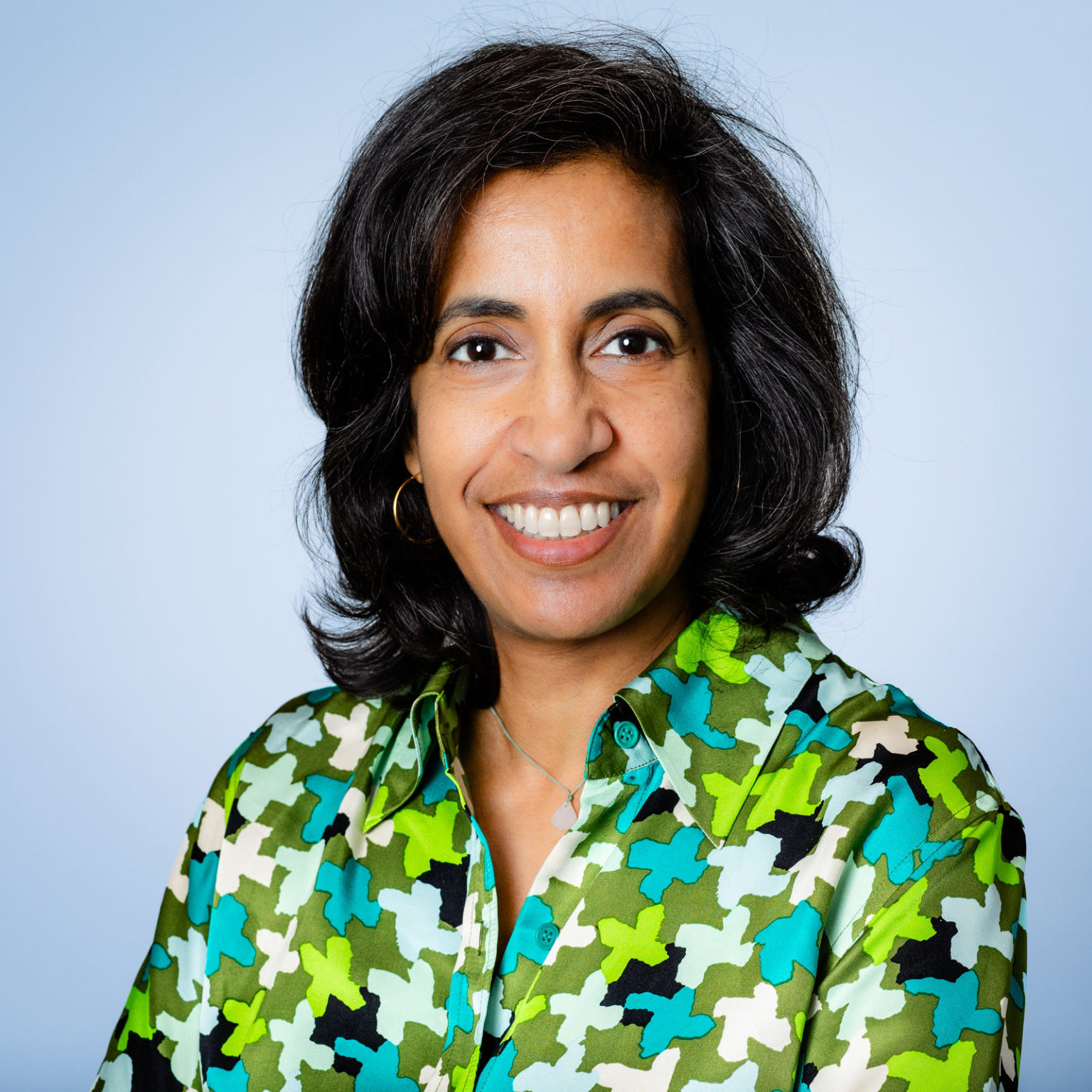 A smiling person with short dark hair wearing a colorful patterned shirt poses against a light blue background.