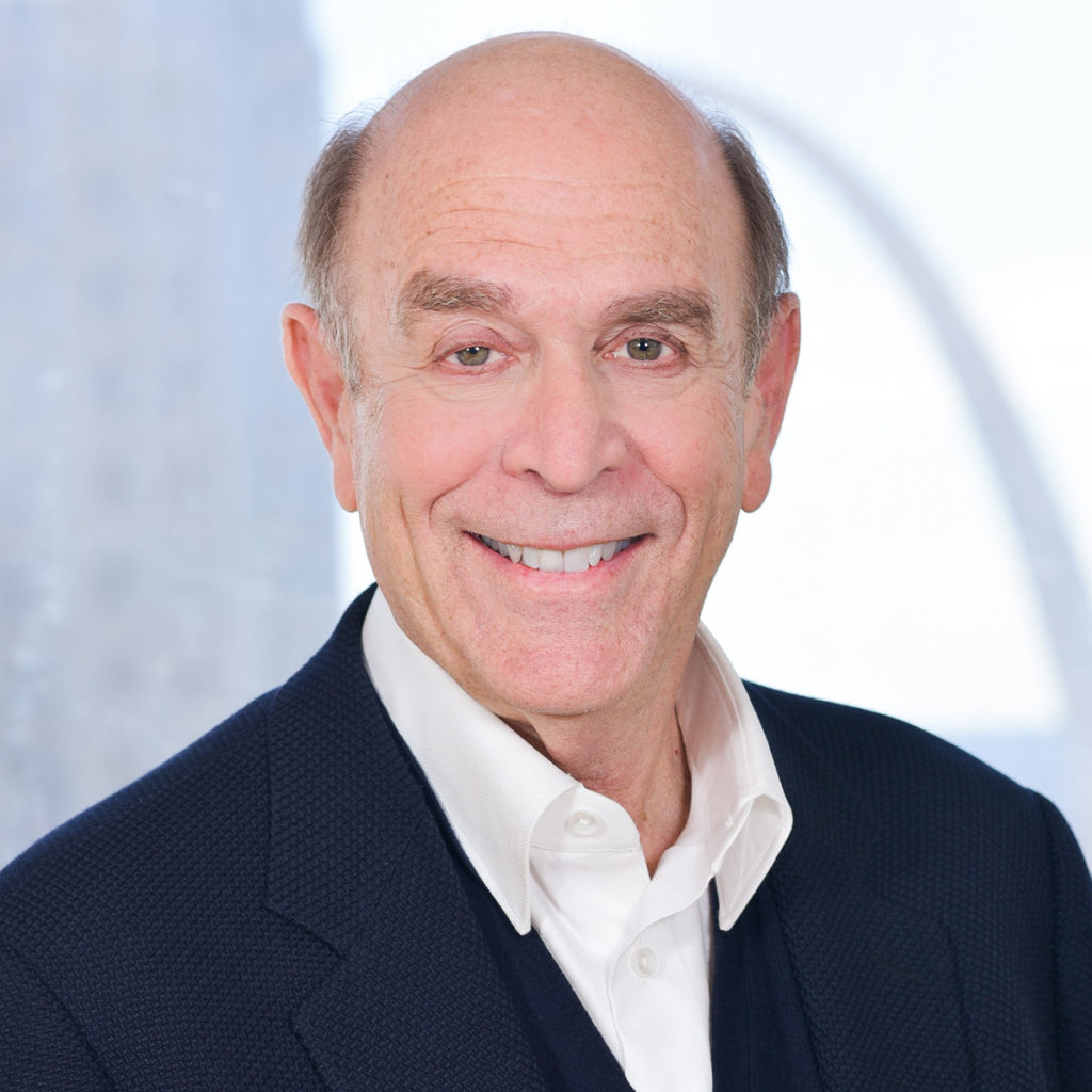 A smiling individual wearing a dark blazer over a white shirt poses against a blurred background.