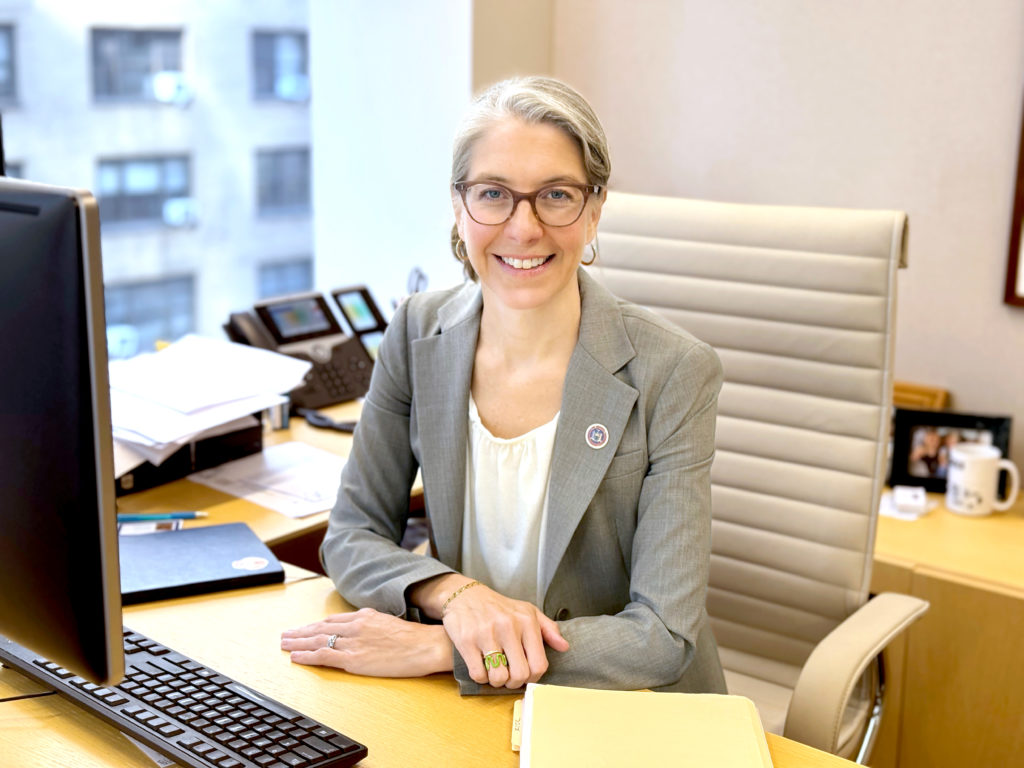 A smiling individual with glasses wearing a gray blazer is seated in an office environment.