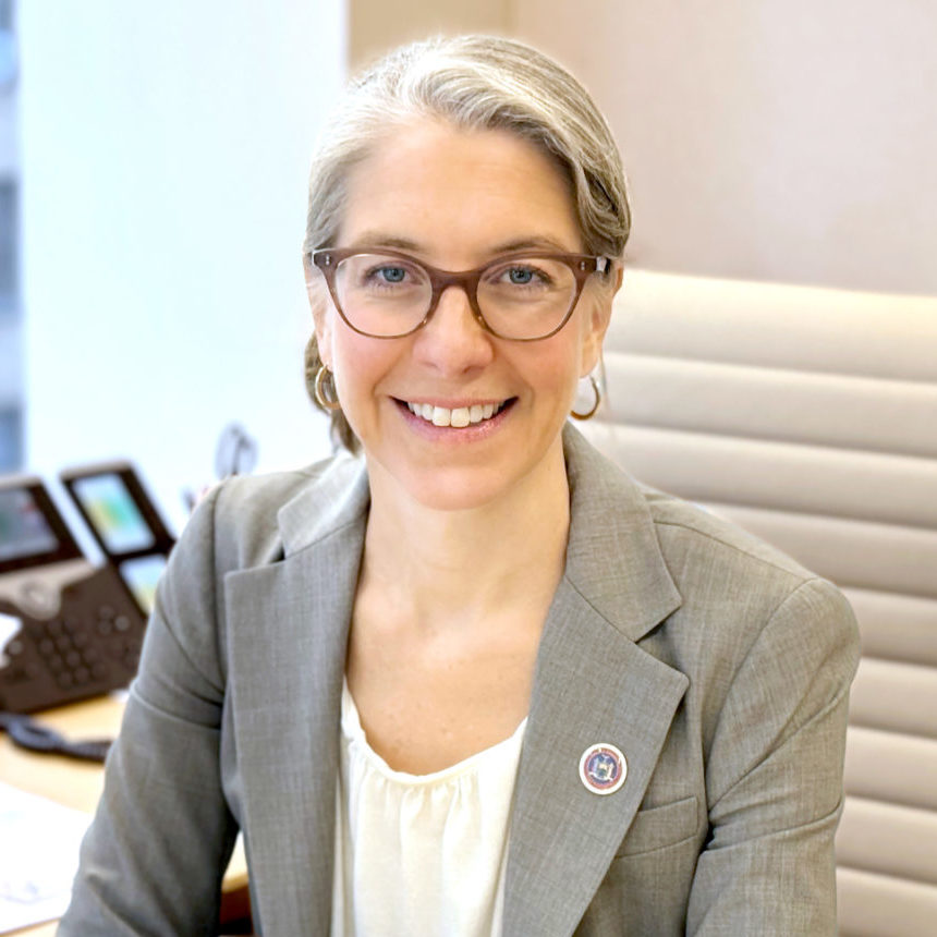 A smiling individual with glasses wearing a gray blazer is seated in an office environment.