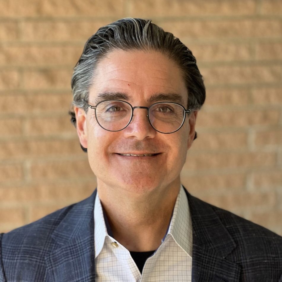 A smiling individual with gray hair and glasses is wearing a suit jacket and a checked shirt against a brick wall backdrop.