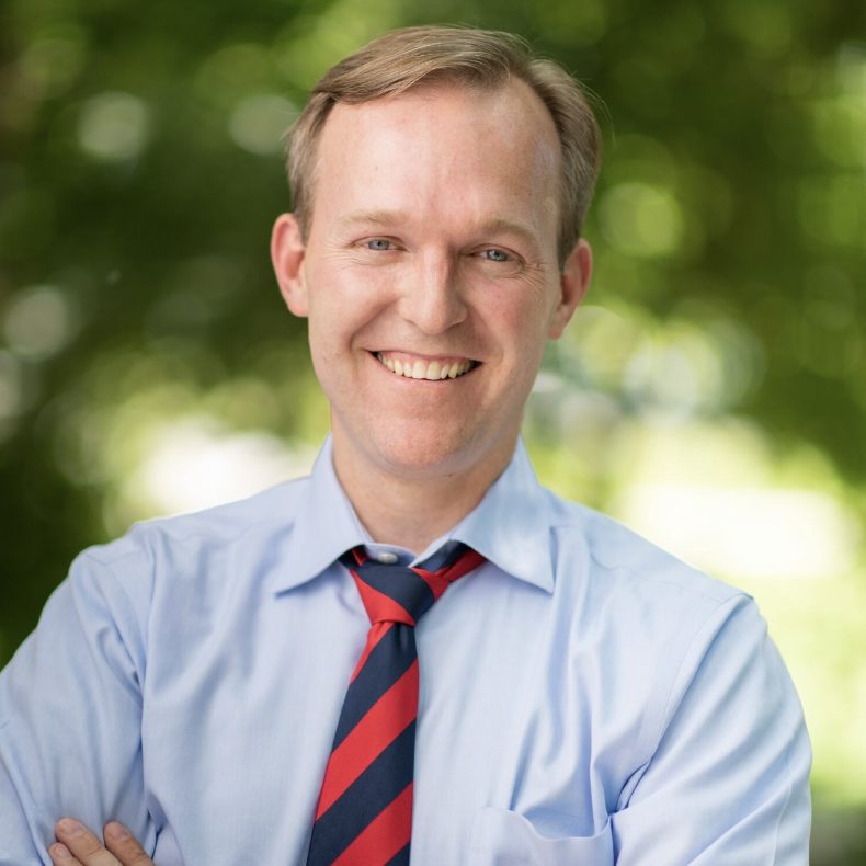 A smiling individual with light hair wears a light blue shirt and a red and blue striped tie against a blurred green background.