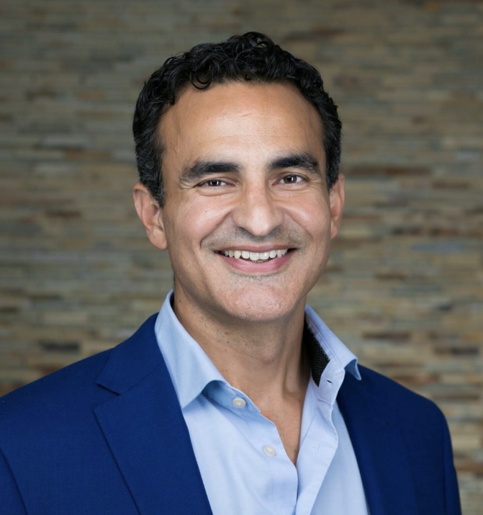 A smiling individual wearing a blue suit jacket over a white shirt against a textured beige background.