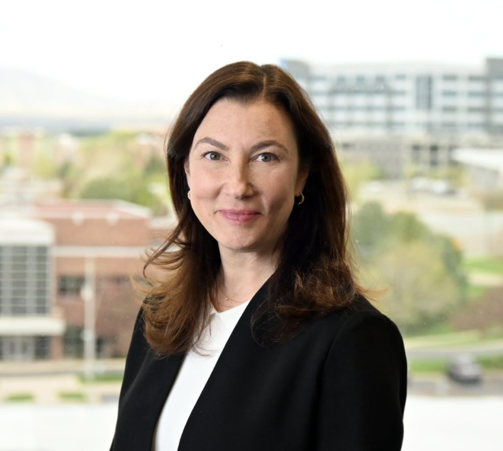 A professional-looking individual with a slight smile stands before a blurred background of urban buildings.