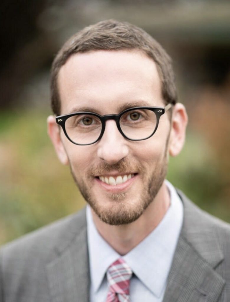 A smiling individual with glasses is wearing a suit and has a neatly trimmed beard.