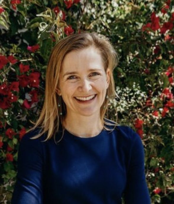 A smiling person wearing a blue top is standing in front of a background with abundant red flowers.