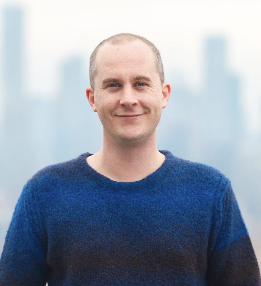 A smiling individual wearing a blue sweater stands in front of a foggy cityscape.