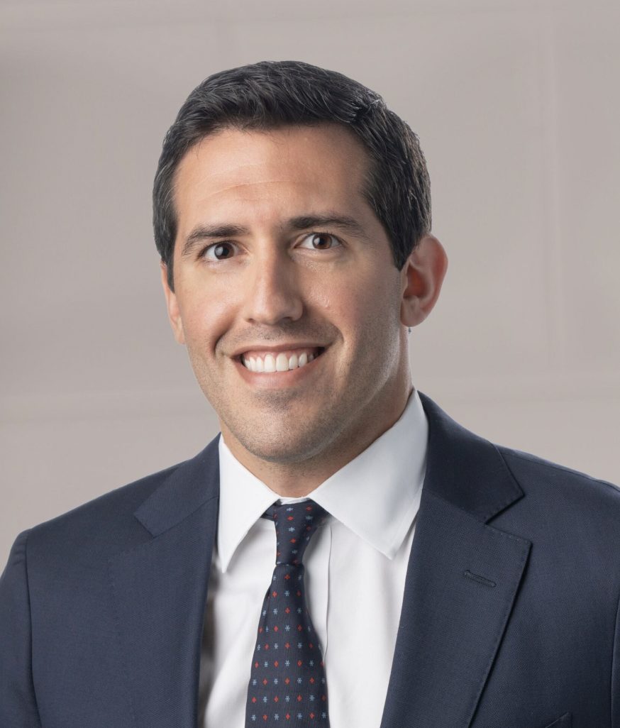 A professional-looking individual is wearing a suit and tie with a pleasant expression against a neutral backdrop.