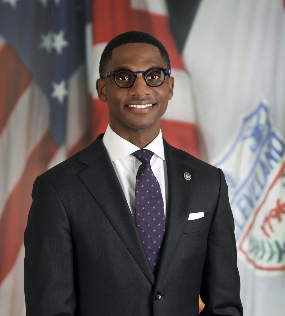 A person wearing glasses and a dark suit stands confidently in front of an American flag and another banner with various insignia.