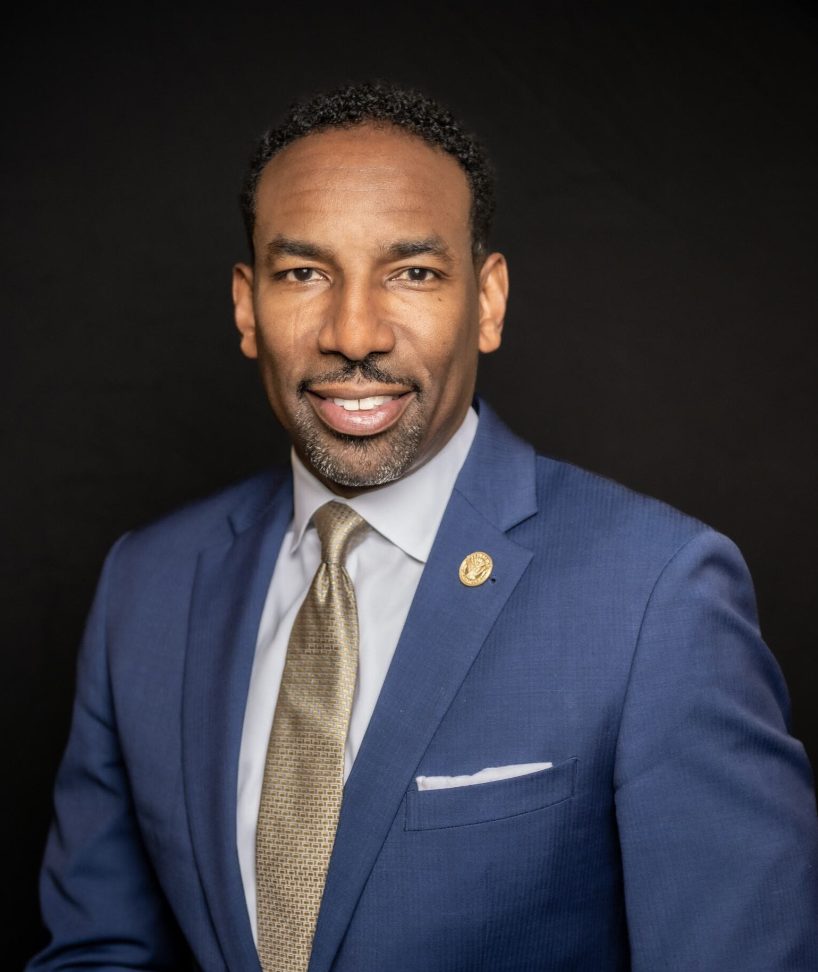 A person is wearing a professional navy blue suit with a tie and has a confident smile.