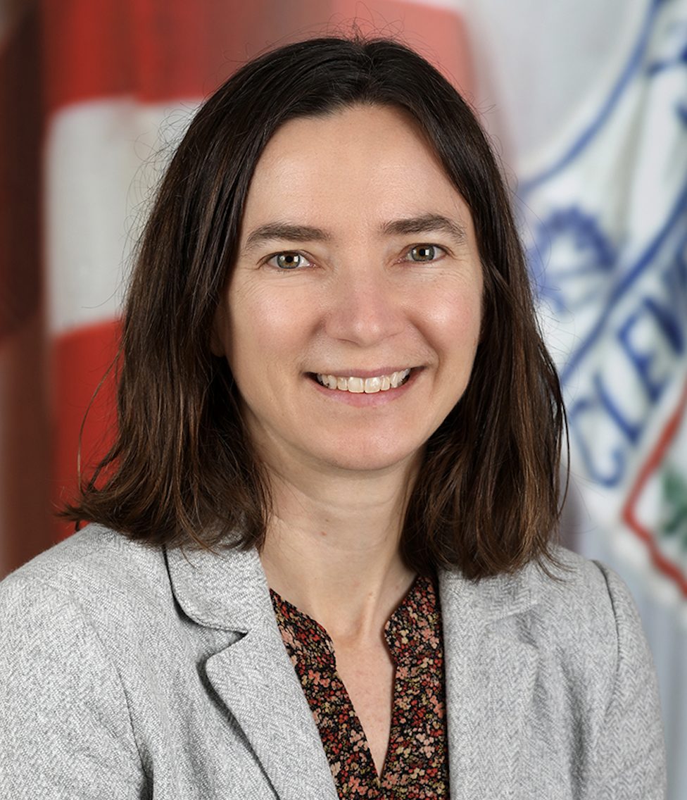 A smiling individual with shoulder-length hair is wearing a gray blazer over a patterned top, with a flag and emblem in the background.