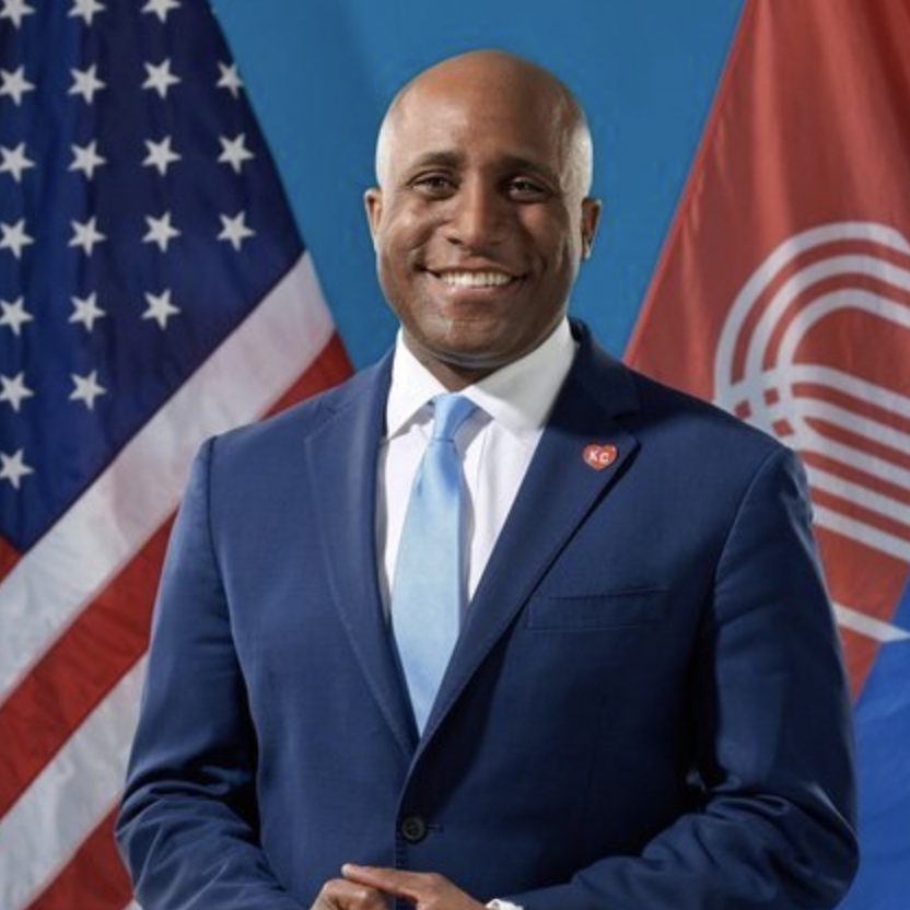 A person in a blue suit stands smiling in front of the United States and Peace Corps flags.