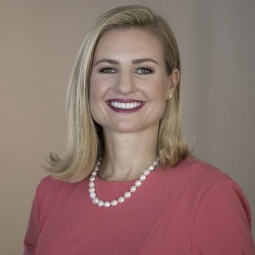 A smiling individual with short blonde hair, wearing a pink top and a pearl necklace, is posing against a neutral backdrop.