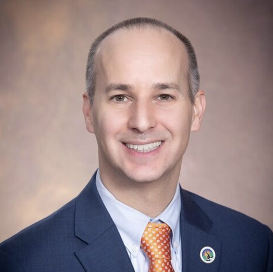 A smiling individual with a shaved head is dressed in a professional navy suit, wearing a patterned orange tie and a circular pin on their lapel.