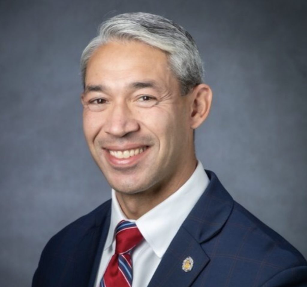 A smiling individual wearing a suit with a red tie and a lapel pin poses for a professional portrait.