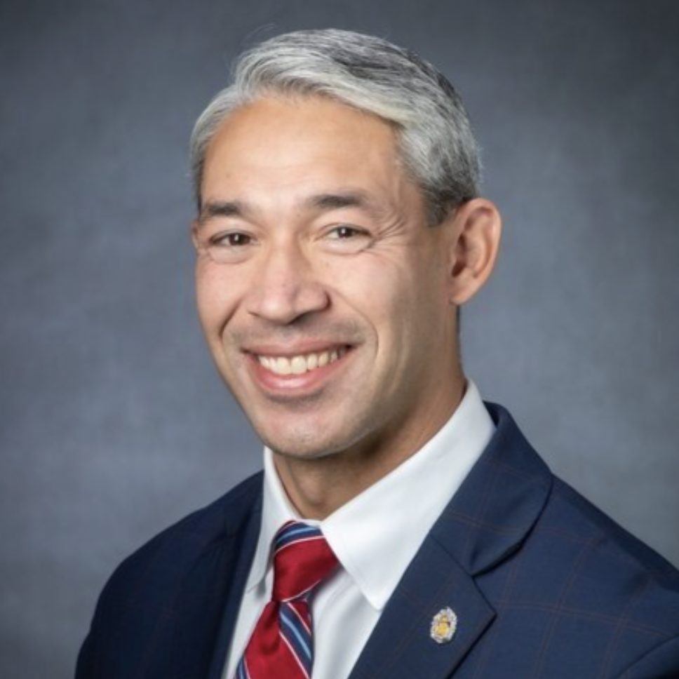 A smiling individual wearing a suit with a red tie and a lapel pin poses for a professional portrait.