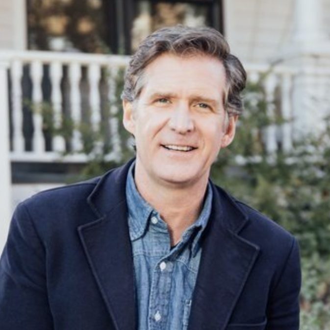 A smiling individual with graying hair is wearing a dark jacket over a blue shirt and stands before a blurred background that suggests a residential setting with white fencing.