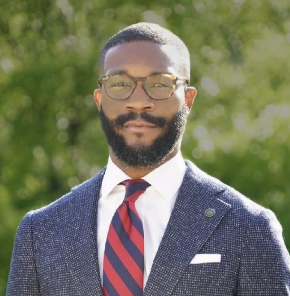 A well-dressed individual with a beard and glasses is wearing a patterned suit, striped tie, and a lapel pin, standing outdoors with trees in the background.