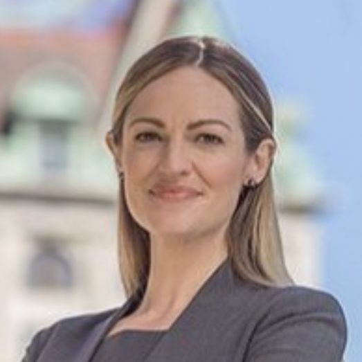 A person is smiling while wearing a formal suit, standing with a blurred background of buildings.