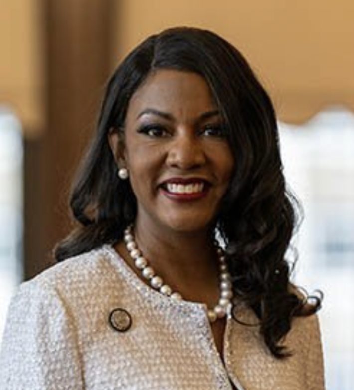 A smiling individual with pearl earrings and a pearl necklace is wearing a light-colored, textured outfit with a round pin on the left side.