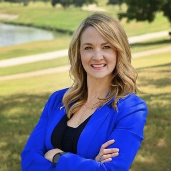 A smiling individual wearing a blue blazer poses confidently with arms crossed in a sunny outdoor setting with a water body and greenery in the background.