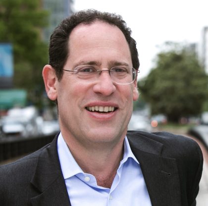 A smiling individual with glasses is captured in a candid moment outdoors, wearing a blue shirt and a dark jacket.