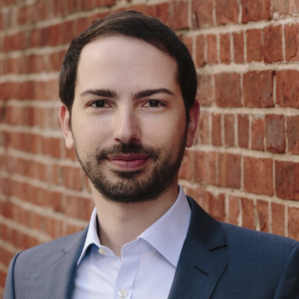 A person with a light stubble is wearing a suit and a blue shirt, posing in front of a brick wall.