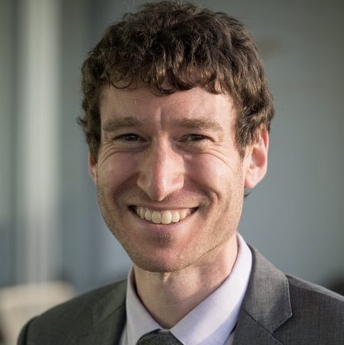 A smiling individual with short curly hair wearing a suit and tie poses for a portrait.