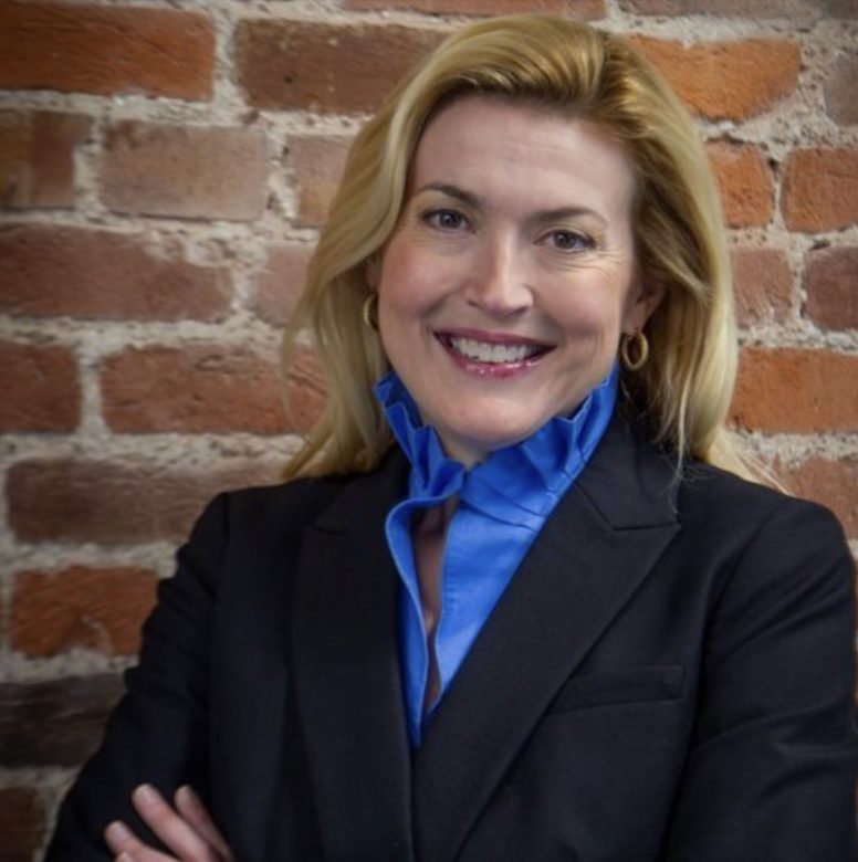 A person is smiling in front of a brick wall, wearing a black blazer and a blue shirt.