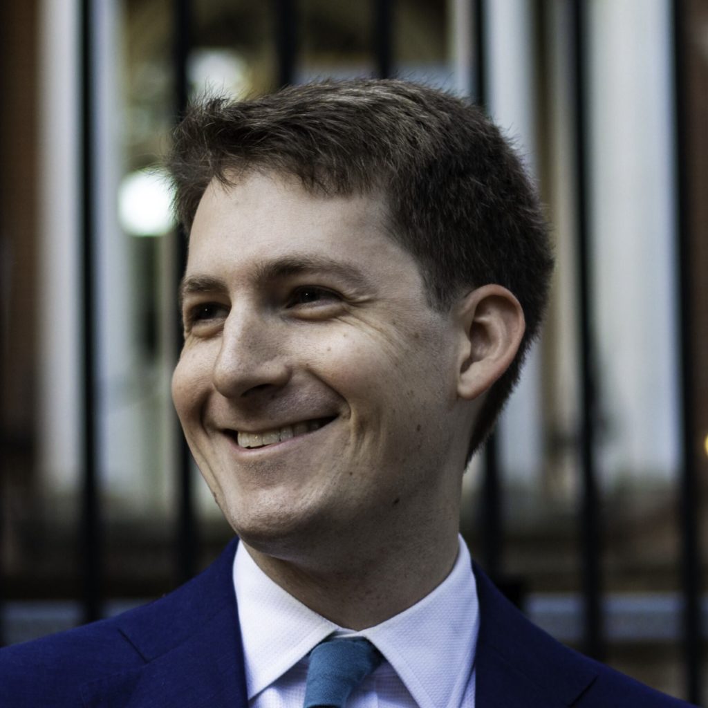 A smiling individual wearing a blue suit and a turquoise tie stands in front of a blurred background.