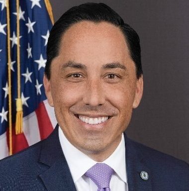 A person is smiling in front of the American flag, dressed in a formal suit with a tie.