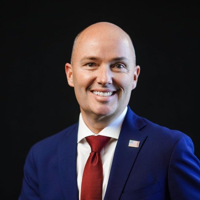 A smiling individual with a shaved head is wearing a dark suit, white shirt, and red tie, against a black background.