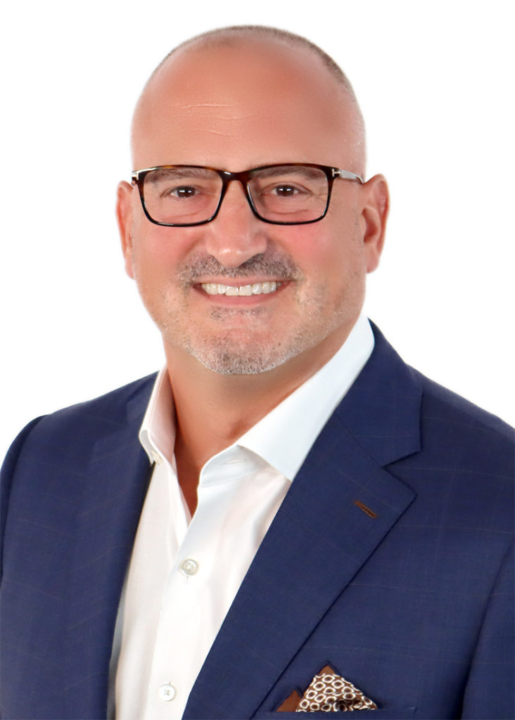 A smiling individual with a shaved head and glasses, wearing a dark suit jacket, poses against a white background.