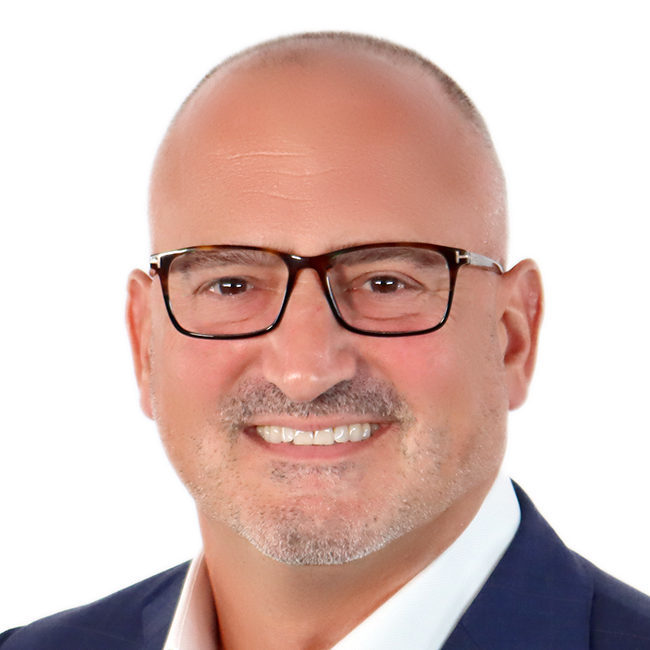 A smiling individual with a shaved head and glasses, wearing a dark suit jacket, poses against a white background.