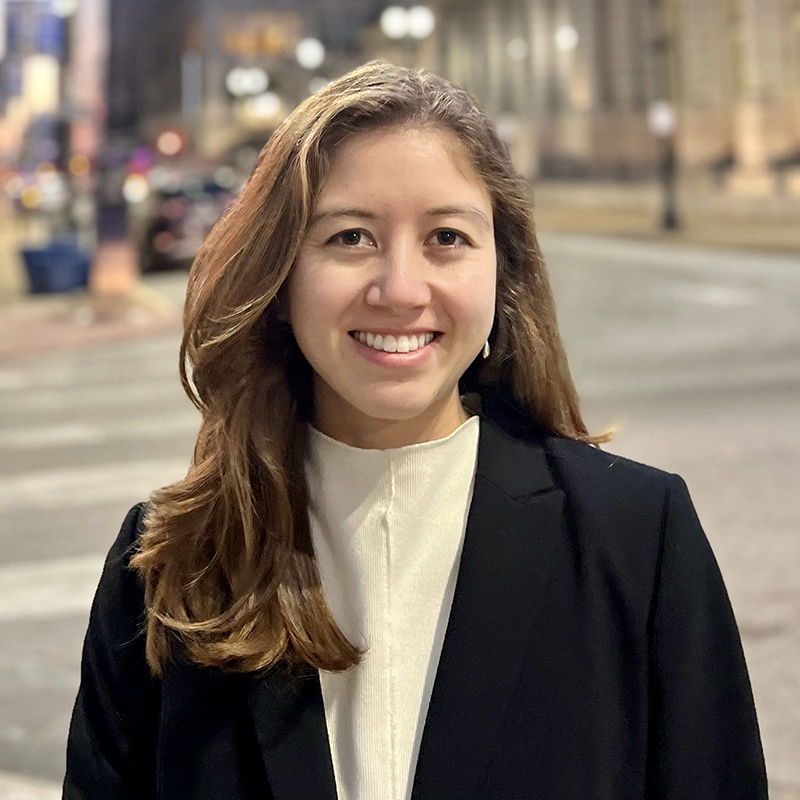 A person is smiling for a portrait with a street scene in the background at dusk or night.