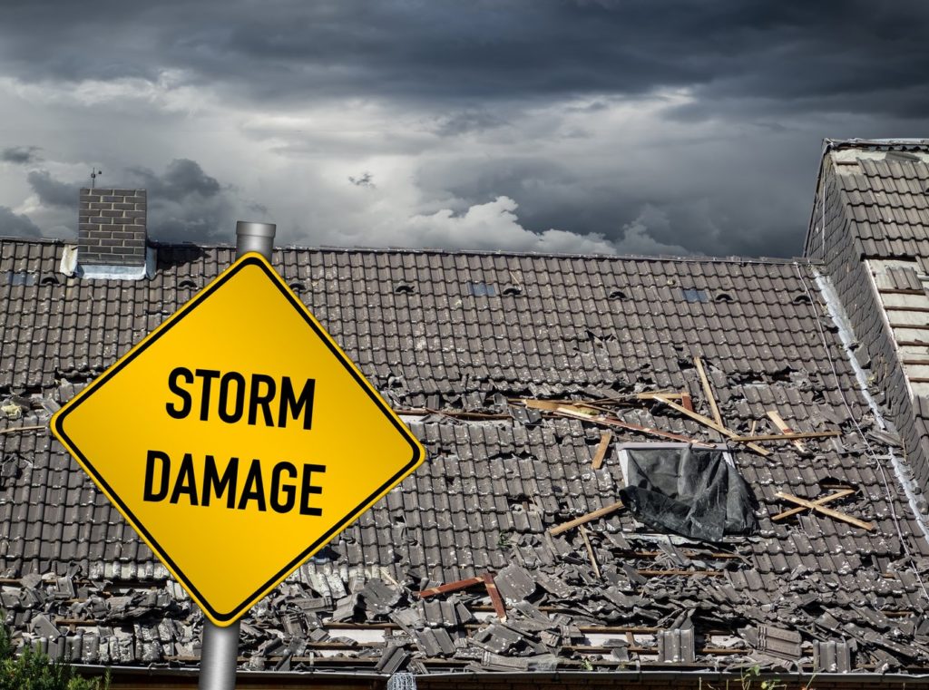 A storm damage warning sign stands in the foreground with a backdrop of a heavily damaged building under overcast skies.