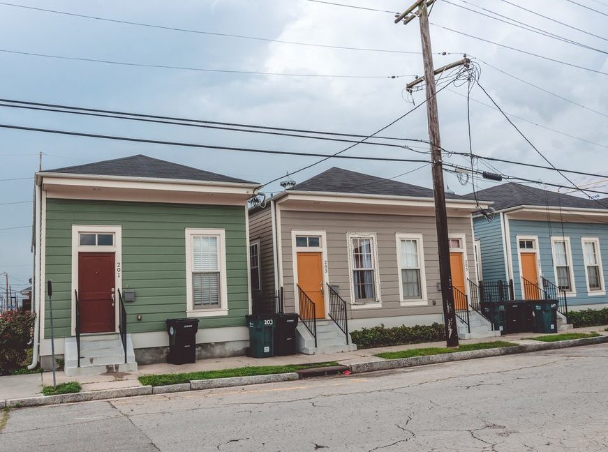 Colorful, identical houses are lined up along a street under an overcast sky with power lines crisscrossing above.