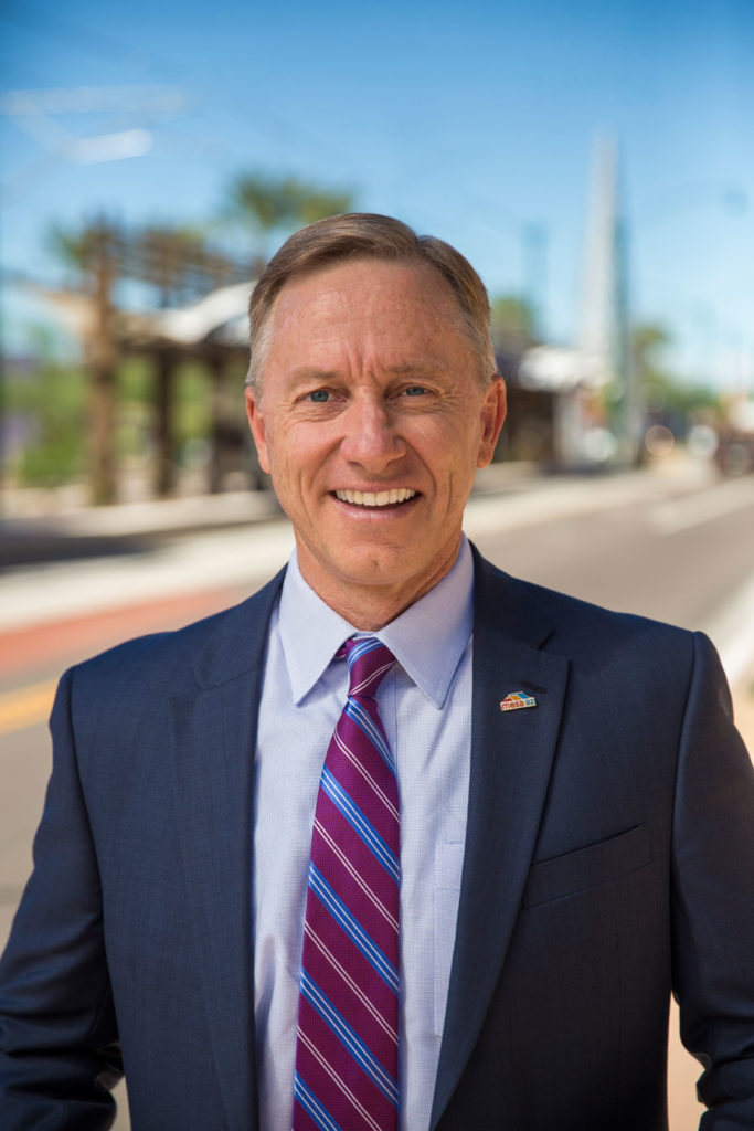 A smiling individual wearing a suit with a tie stands outdoors with a street and buildings in the background.