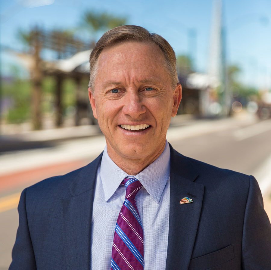 A smiling individual wearing a suit with a tie stands outdoors with a street and buildings in the background.