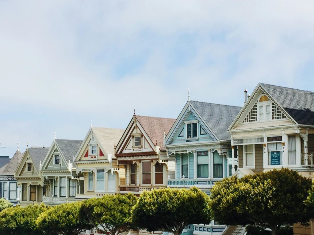 Victorian houses stand in a row under a cloudy sky, showcasing ornate architectural details and a variety of pastel colors.