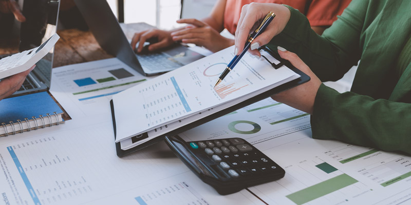 A person is analyzing financial charts and using a calculator with a laptop in the background.