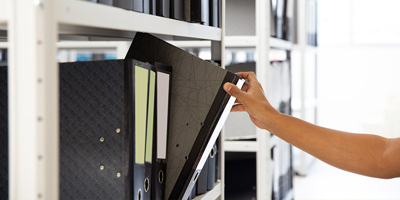 A hand is pulling a file binder from a shelf among several other binders.